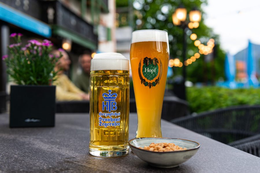 Im Biergarten des Hotel Terofal in Schliersee sieht man ein Weißbier und ein Helles. Im Hintergrund sieht man das Hotel, unscharfe Lichter und zwei Gäste die ihren Tag genießen.