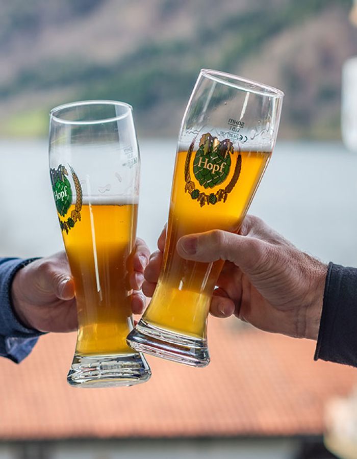 Two guests in the beer garden at the Terofal in Schliersee clinking full wheat beer glasses, with Lake Schliersee visible in the background