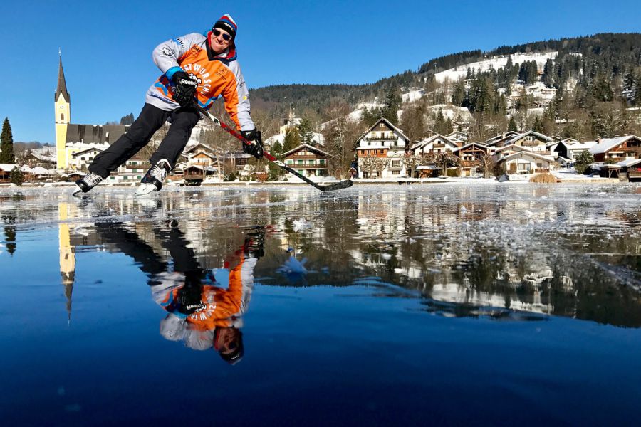 Ein Mann spielt auf dem zugefrohrenen Schliersee Eishockey, im Hintergrund sind die Ferienwohnungen der Seeheimat in Schliersee zu sehen