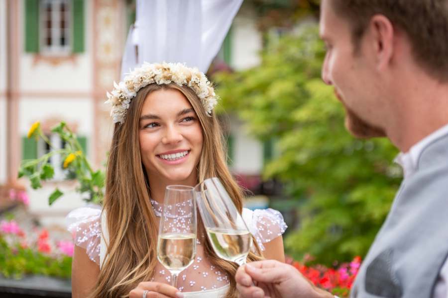 Glückliche Braut stößt mit einem Glas Sekt an und lächelt in die Kamera bei ihrer Hochzeit am Schliersee